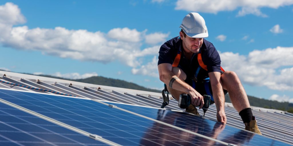 Solar Audit Compliance Check 5 A worker wearing a white helmet crouches on a rooftop in Brisbane, installing solar panels with a power drill under the bright sunshine typical of the Sunshine Coast, with blue sky and clouds overhead.