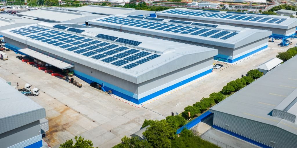 Aerial view of large industrial warehouses with solar panels on the roofs near Brisbane, surrounded by lorries, loading bays, and trees, on a bright and clear day.