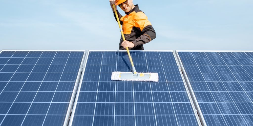 Solar Panel Cleaning 2 A worker in an orange safety vest and helmet cleans large solar panels with a long-handled mop under the clear blue sky on the Sunshine Coast.