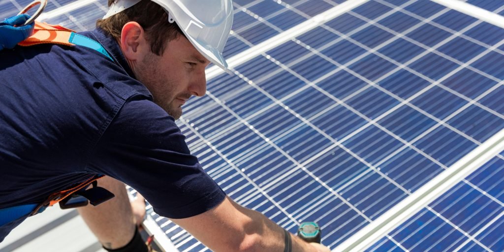 Solar Health Checks 1 A person wearing a white hard hat and safety gear installs or inspects solar panels on a rooftop in Brisbane, working with tools and focusing on the task.