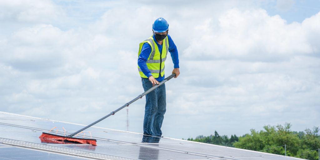 Maintenance Plan 3 A worker in a blue hard hat, safety vest, and mask uses a long brush to clean solar panels on a rooftop under a cloudy Brisbane sky. Trees are visible in the background.