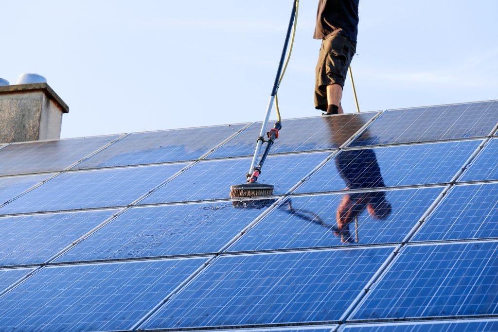 A person in Brisbane stands on a roof using a long-handled brush to clean solar panels, with the blue sky reflected on their shiny surfaces.