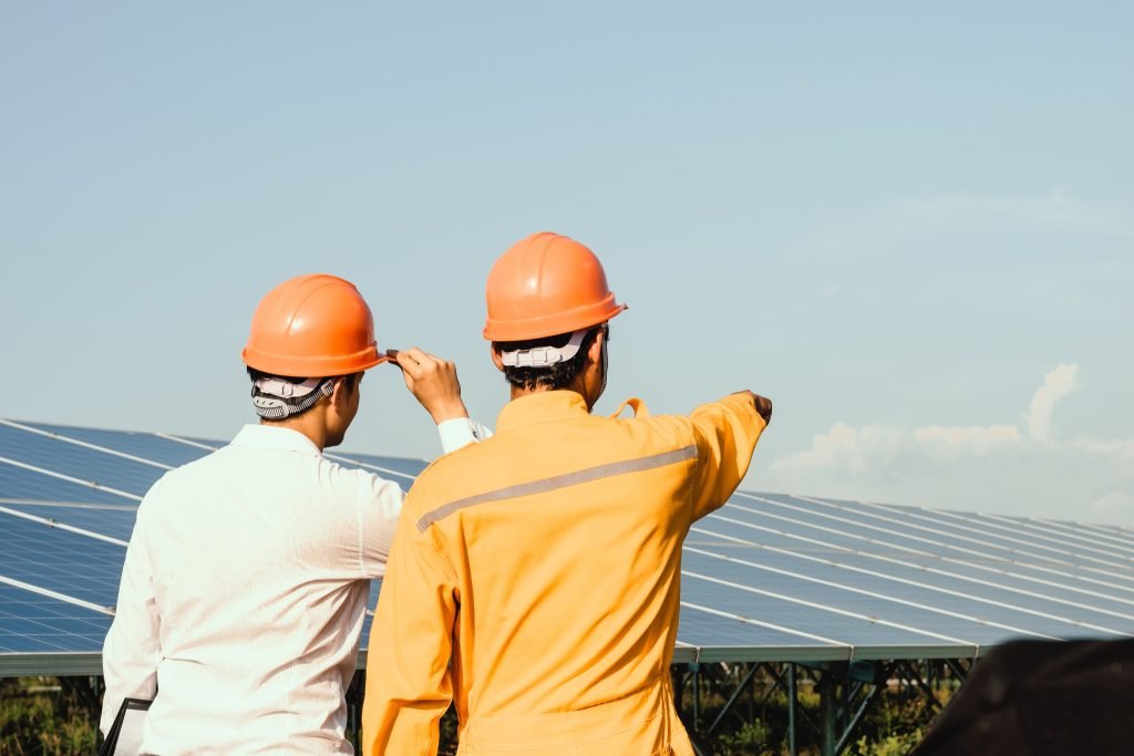 Sunshine Coast Solar System Upgrade 1 Two workers wearing orange safety helmets stand near large solar panels outdoors. One worker, dressed in yellow, points ahead as they both look towards the solar array under the clear blue sky on the Sunshine Coast near Brisbane.