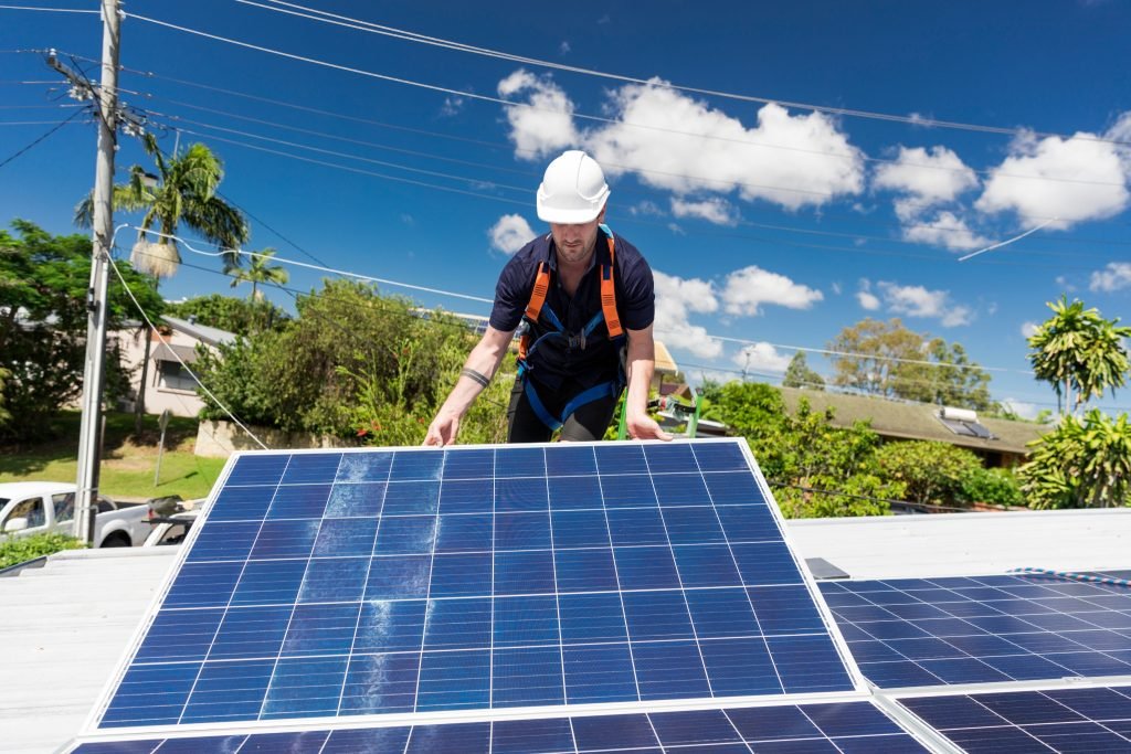 A worker wearing a white hard hat and safety harness installs solar panels on a rooftop under a bright blue sky, with scattered clouds and trees in the background, bringing renewable energy to Brisbane and the Sunshine Coast.