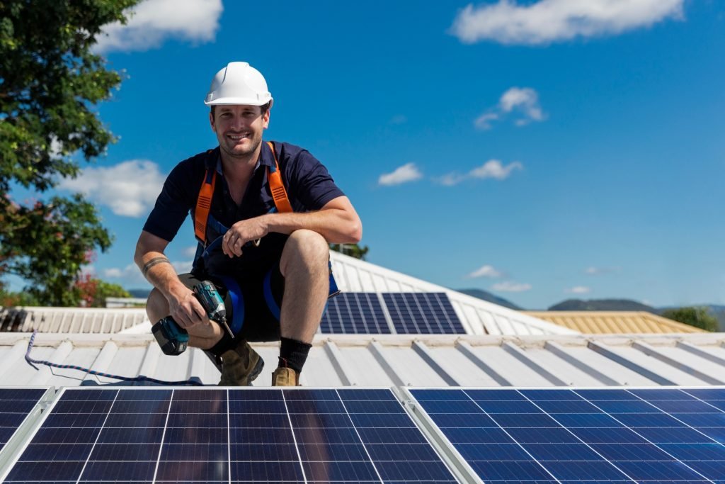 Design & Install 2 A man wearing a white hard hat and safety harness kneels on a roof in Brisbane, smiling with a power drill in hand beside solar panels under a bright blue sky.