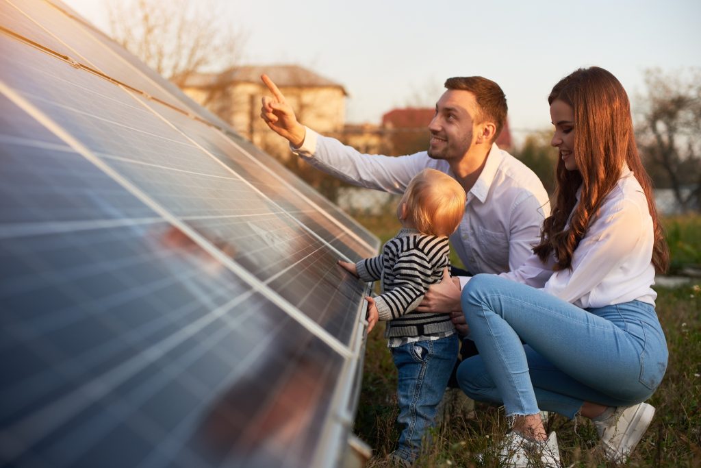 A man, woman, and young child kneel outdoors on the Sunshine Coast, examining and touching solar panels together under bright sunlight, with houses visible in the background.