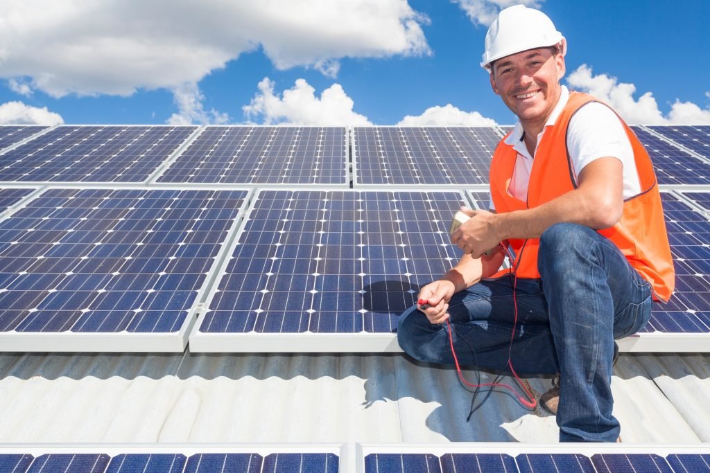 Commercial Solar Health Checks 1 A smiling worker in a white hard hat and orange safety vest kneels on a roof beside solar panels, working with electrical wires under the bright Brisbane sky. Blue sky with clouds is visible in the background.