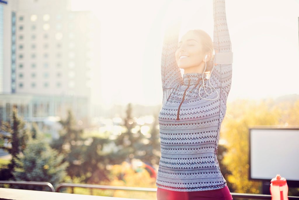 Woman in sportswear stretches outdoors with arms raised, smiling in the sunlight. She has earphones in and a fitness tracker on her arm, enjoying a sunny day near Brisbane with trees and tall buildings in the background.