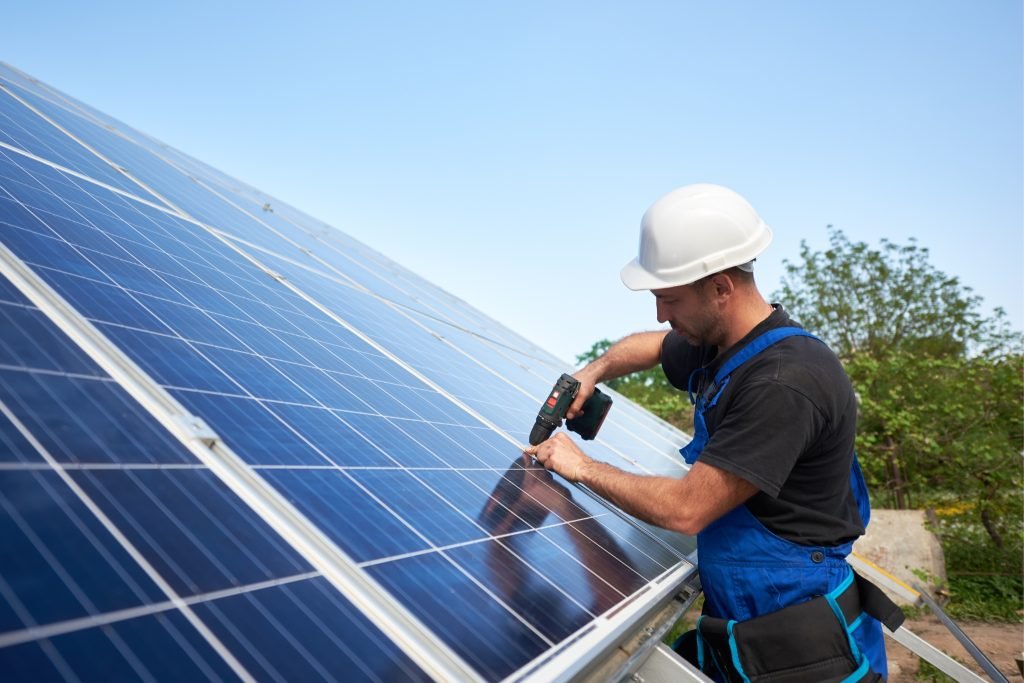The Advantages of Solar Design & Installation for Your Home or Business 1 A worker in a white hard hat and blue overalls uses a power drill to install solar panels outdoors on a sunny day, perfect for solar projects in Brisbane or the Sunshine Coast, with trees and a clear sky in the background.