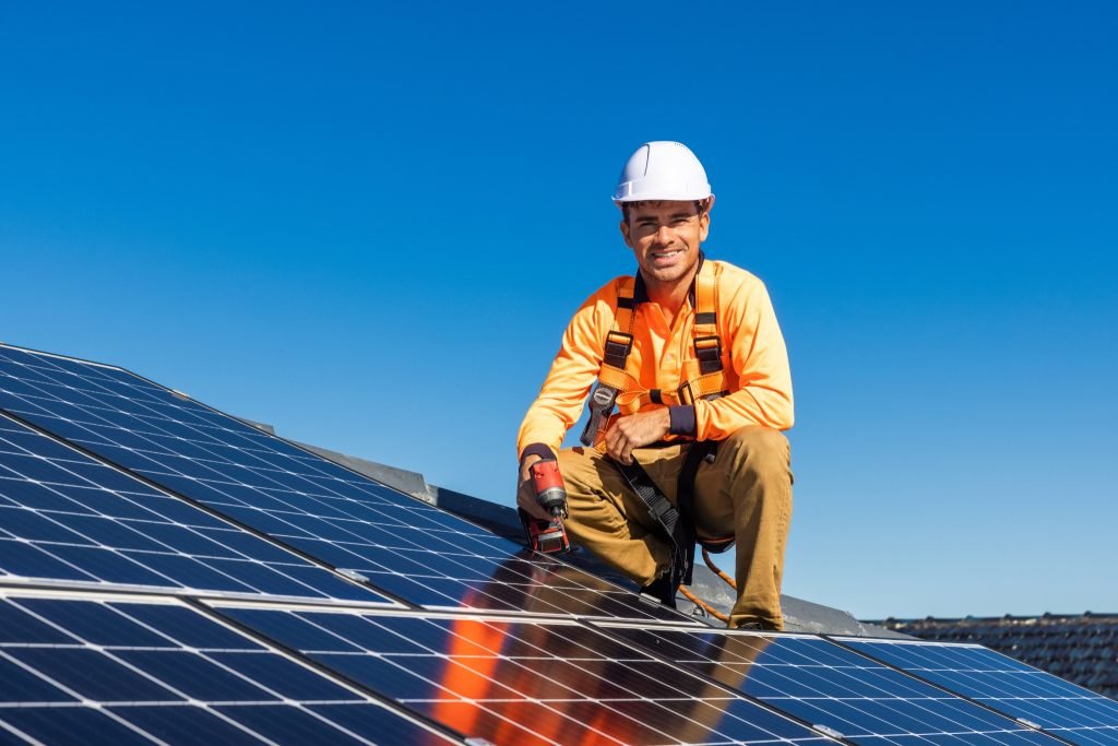 Commercial Solar Services 3 A worker in a hard hat and safety harness kneels on a rooftop, installing solar panels under the clear Brisbane sky, holding a power drill and smiling at the camera.