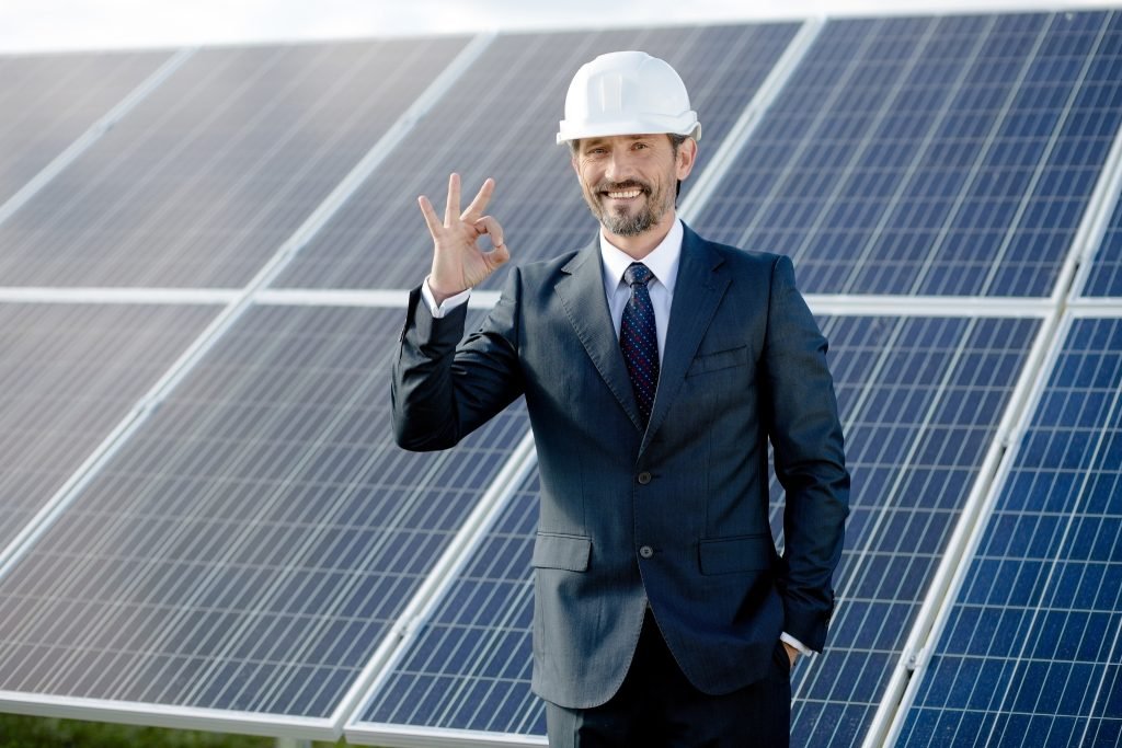A man in a suit and white hard hat stands in front of solar panels, smiling and making an "OK" hand gesture, showcasing the bright future of solar energy on the Sunshine Coast.