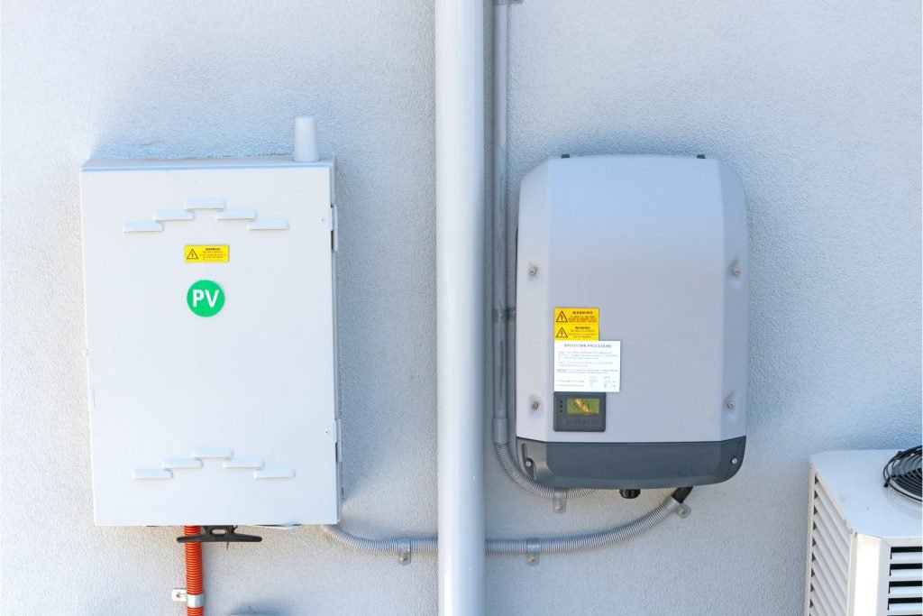Solar Health Checks 2 Two electrical boxes are mounted on a light grey wall. The left box, labelled with a green PV solar sticker, and the right box, marked with warning labels, are connected by a white pipe—common in Brisbane and Sunshine Coast installations.