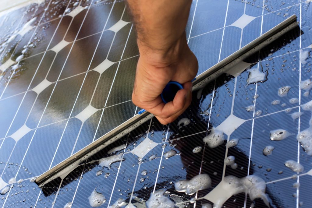 A hand uses a squeegee to clean a solar panel covered with soap and water, ensuring the panel’s surface is clear for optimal sunlight absorption in Brisbane and the Sunshine Coast.