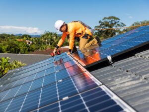 Solar System Health Checks & Performance Audits Strathpine 1 A worker in safety gear installs solar panels on a rooftop under a clear blue sky in Brisbane, using a power drill. Trees and houses are visible in the background.
