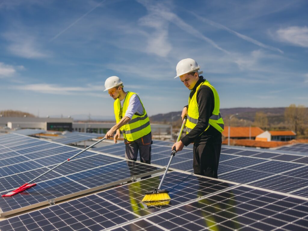 Two workers in safety helmets and high-visibility vests clean solar panels on a Brisbane rooftop with brushes on a sunny day, blue sky above and distant buildings in the background.