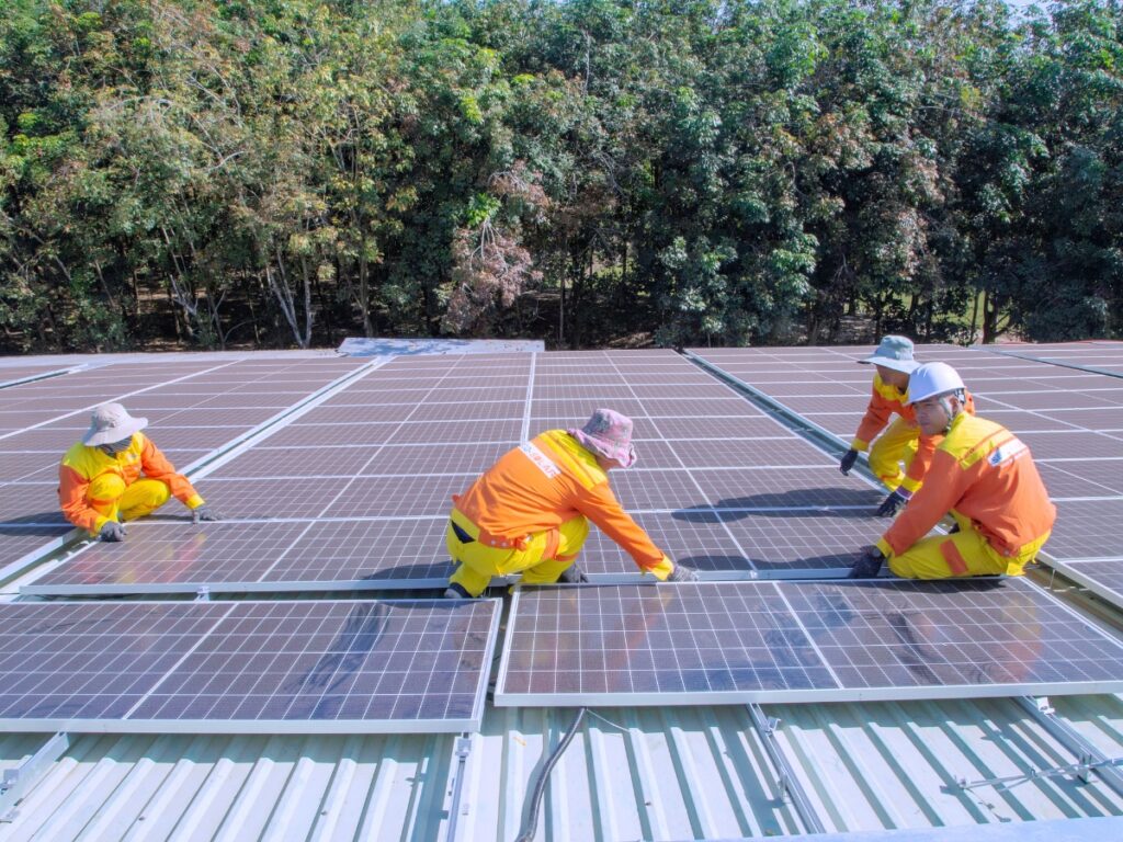 Four workers in orange and yellow uniforms are installing solar panels on a rooftop, bringing clean energy to the Sunshine Coast. Dense green trees provide a scenic backdrop as they work under clear skies.