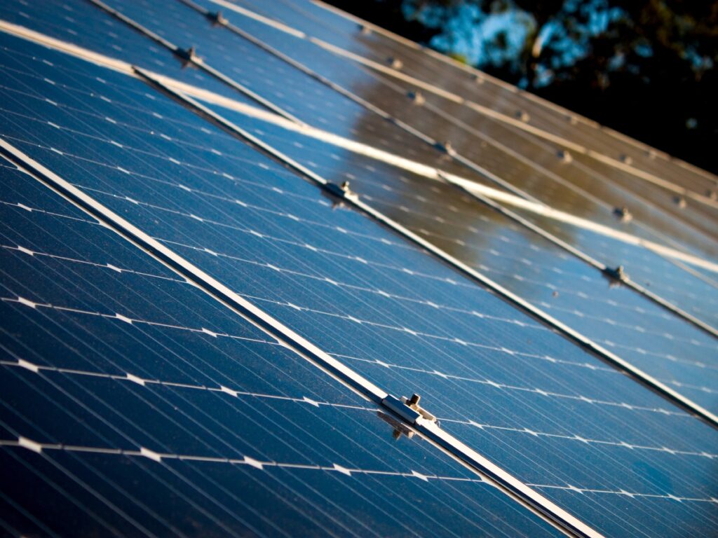 Close-up view of several blue solar panels angled towards the sky in Brisbane, reflecting sunlight, with blurred trees and a blue sky in the background.