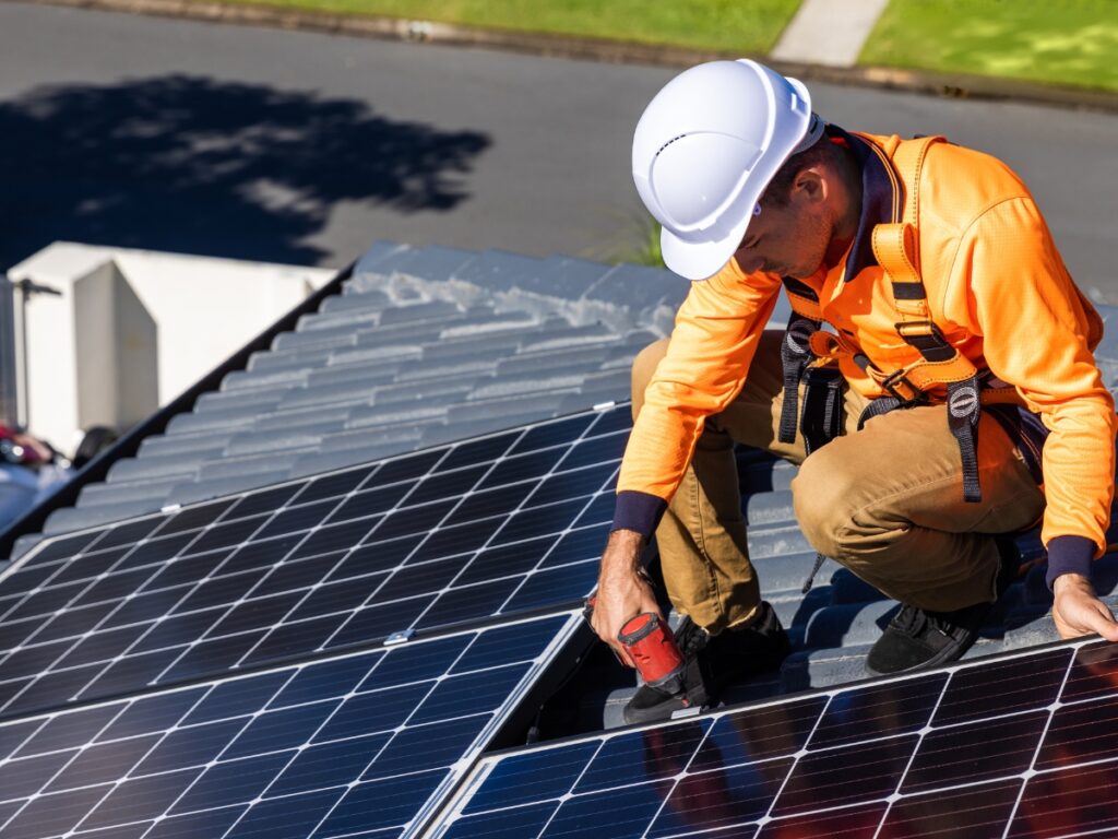 A worker in a safety helmet and orange jacket installs solar panels on a residential rooftop in Brisbane using a power drill, with a street and green grass in the background.