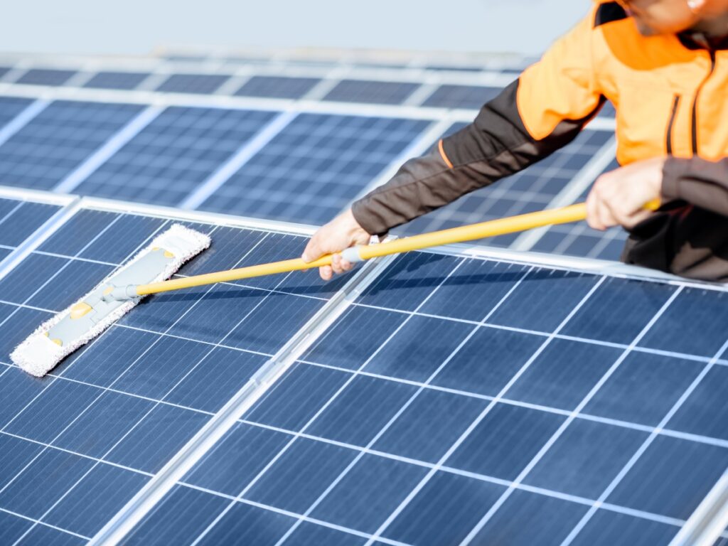 Solar Panel Maintenance on the Sunshine Coast: Pro Tips vs. DIY Risks 9 A person in an orange jacket uses a yellow-handled mop to clean the surface of large solar panels outdoors, maintaining solar efficiency under the bright Sunshine Coast sun.