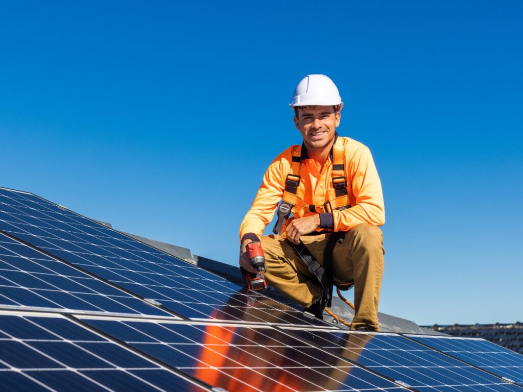 Solar Power Sunshine Coast 101: How It Works & Benefits | Limitless Solar 10 A worker in a hard hat and high-visibility clothing kneels on a rooftop in Brisbane, installing solar panels with a drill under a clear blue sky.