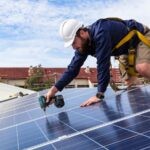 Best Solar Solutions: Noosa to North Brisbane Guide 1 A worker wearing a safety harness and helmet uses a power drill to install solar panels on a Brisbane rooftop under a partly cloudy sky.
